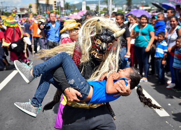 Человек в маске мифологического существа держит на руках девочку во время парада в Сан-Сальвадор - Sputnik Южная Осетия